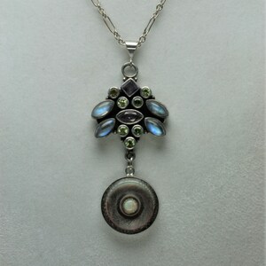 May include: A silver necklace with a pendant featuring a cluster of moonstone, amethyst, and peridot gemstones. The pendant is attached to a round, silver charm with a white opal gemstone in the center.
