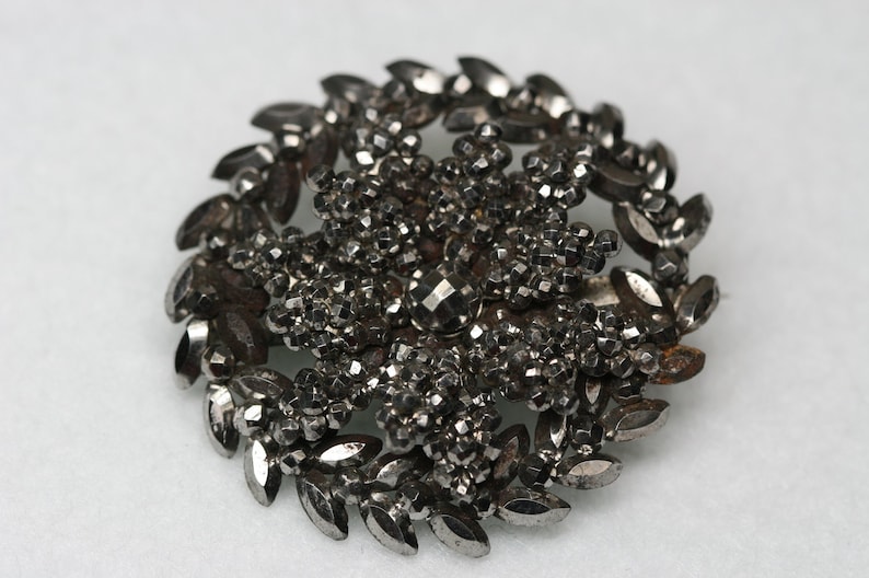 Pu&ograve; includere: Un fermaglio argentato con un designo circolare. Il centro del fermaglio &egrave; un gruppo di piccole perline sfaccettate, circondato da un anello di perline pi&ugrave; grandi a forma di foglia.