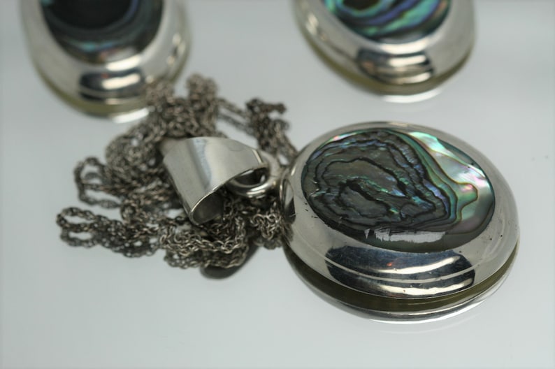 May include: A silver chain necklace with a pendant featuring a round abalone shell with iridescent green, blue, and black colors.