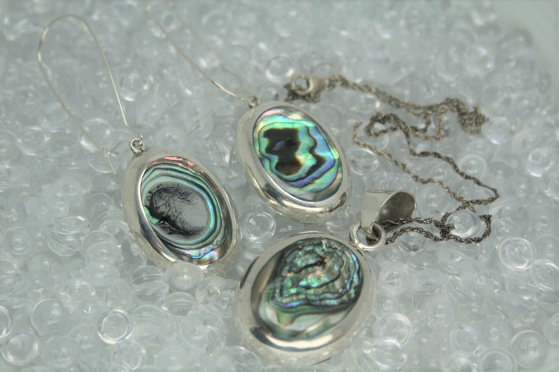 May include: A set of three silver oval pendants with iridescent abalone shell inlays. The pendants are on a silver chain and a pair of silver earring hooks.