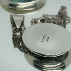 May include: Silver pendant with a round, silver charm. The charm has the word "MEXICO" and the number "925" engraved on the back.