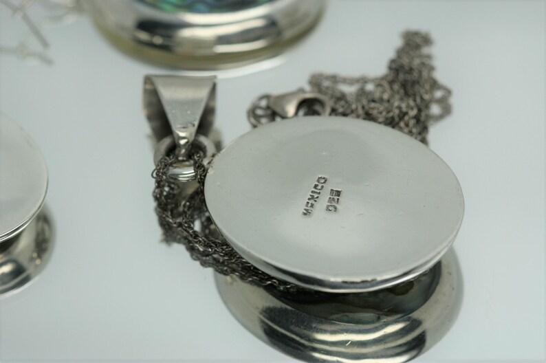 May include: Silver pendant with a round, silver charm. The charm has the word "MEXICO" and the number "925" engraved on the back.