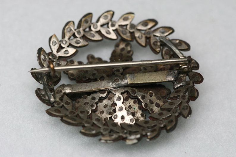 Pu&ograve; includere: Un fermaglio argentato con un design a forma di stella circondato da una ghirlanda di foglie. Il fermaglio ha una chiusura a spilla e gancio sul retro.