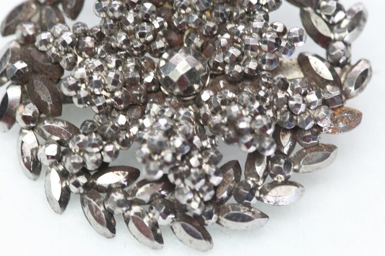 Pu&ograve; includere: Un fermaglio argentato con una pietra centrale sfaccettata a forma di diamante circondata da piccole perle rotonde. Il fermaglio ha la forma di un fiore con foglie fatte delle stesse perle.