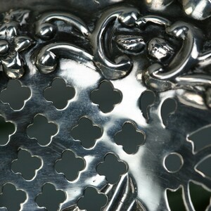 Puede incluir: Primer plano de un colador de plata con un dise&ntilde;o floral y una superficie perforada. El colador tiene un acabado met&aacute;lico brillante.