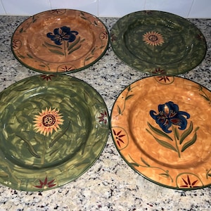 May include: Four decorative ceramic plates with floral designs. Two plates have a brown background with a blue flower, and two have a green background with a yellow sunflower. Each plate has a green rim and measures approximately 25 cm in diameter.