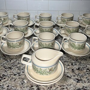Op de afbeelding: Een set crèmekleurige kopjes en schotels met een bloemen- en vlinderontwerp, samen met een melkkan. Elke kop heeft een oor en een blauwe rand. De set bevat 16 kopjes en schotels.