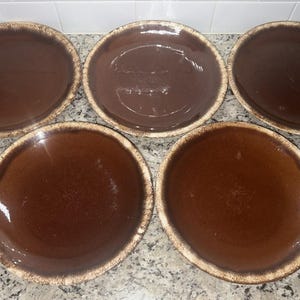 Könnte beinhalten: Fünf braune Keramikplatten mit glänzendem Finish und einem strukturierten, hellfarbigen Rand. Die Teller sind auf einer gesprenkelten Arbeitsplatte angeordnet. Die Teller dienen wahrscheinlich zum Servieren von Speisen.