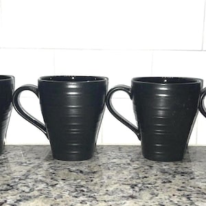 May include: Four black ceramic mugs with a ribbed design are arranged in a row. Each mug has a curved handle and a slightly tapered shape. The mugs are sitting on a speckled countertop.