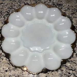 Pode incluir: Um prato branco, com bordas onduladas, para servir ovos com uma borda dourada. O prato tem uma área circular central rodeada por 12 compartimentos individuais em forma de ovo. A borda tem uma borda decorativa ondulada.