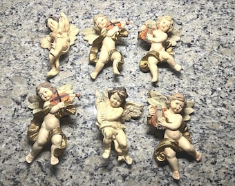Puede incluir: Seis figuras de querubines de color crema con detalles dorados. Algunos querubines sostienen instrumentos musicales, como un violín y un arpa. Los querubines tienen alas detalladas y están posados en varias posiciones. Las figuras están sobre una superficie gris moteada.
