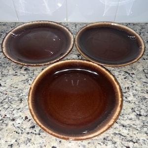 Peut inclure: Trois assiettes en céramique marron avec une finition brillante et un bord marron clair. Les assiettes sont disposées sur un comptoir moucheté. Les assiettes sont probablement destinées à servir des aliments.