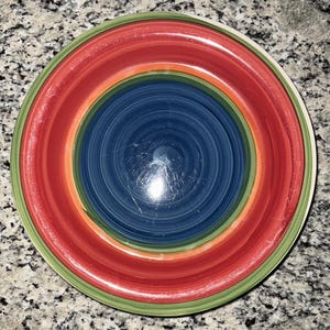 May include: A colorful ceramic plate with concentric rings of color. The center is dark blue, surrounded by orange, red, and green bands. The plate has a glossy finish and is suitable for serving food.