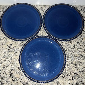 May include: Three round, navy blue ceramic plates. Each plate has a dark brown and light blue striped border. The plates are arranged in a triangular pattern on a speckled countertop.