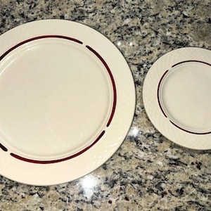 May include: Two round off-white ceramic plates of different sizes, each featuring a thin, dark red circular band accent and a subtle, embossed diagonal cross-hatch pattern along the outer rim of the dinnerware.