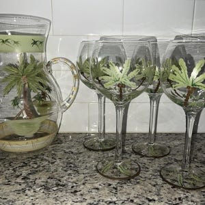 May include: A clear glass pitcher and four wine glasses with a tropical palm tree design. The pitcher has a handle and a green band at the top. The glasses have long stems and a gold rim.