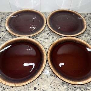 May include: A set of four round ceramic plates featuring a deep, glossy dark brown center that transitions into a lighter, textured tan rim. Each plate has a smooth, reflective surface with a distinct color gradient from the dark interior to the edge.