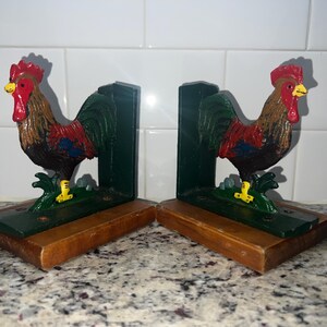 May include: A pair of rooster-shaped bookends. Each bookend features a detailed, colorful rooster with red comb and wattle, yellow beak, and green and brown feathers. The roosters stand on a green base with a wooden bottom.