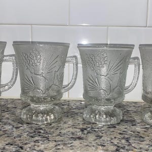 Puede incluir: Juego de cuatro tazas de vidrio transparente con un diseño de piña. Las tazas tienen una base con pie y un asa.