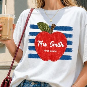 Op de afbeelding: Een wit T-shirt met korte mouwen voorzien van een afbeelding van een rode appel met een groen blaadje over horizontale blauwe penseelstreken. De appel bevat de witte tekst "Mrs. Smith" en "First Grade" in een script en schreefloos lettertype.