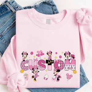 A pink t-shirt with Minnie Mouse and other Minnie Mouse-themed designs, along with a pair of blue jeans, are laid out on a white surface.