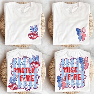 May include: White t-shirts with patriotic designs. One shirt features a peace sign with red, white, and blue stripes and stars. Another has a bow with similar colors. Two shirts have the text "Little Mister Fire Cracker" and "Little Miss Fire Cracker".