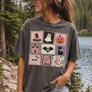 May include: A charcoal gray short-sleeve t-shirt featuring a grid of nine square patches with Halloween-themed illustrations including a witch hat, ghost, pumpkin, haunted house, bat, black cat, skull, spellbook, and crescent moon in pink and black.