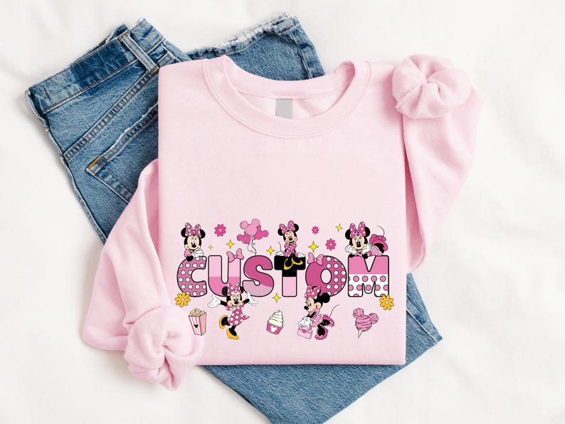 A pink t-shirt with Minnie Mouse and other Minnie Mouse-themed designs, along with a pair of blue jeans, are laid out on a white surface.