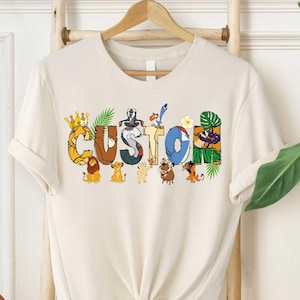May include: A white t-shirt with a graphic of the word "CUSTOM" made up of characters from the Lion King movie. The characters include Simba, Nala, Timon, Pumbaa, and Mufasa.