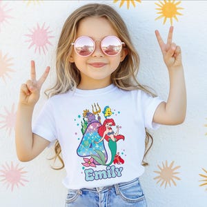 A young girl wearing a white t-shirt with a mermaid design and sunglasses is making a peace sign with her hands.