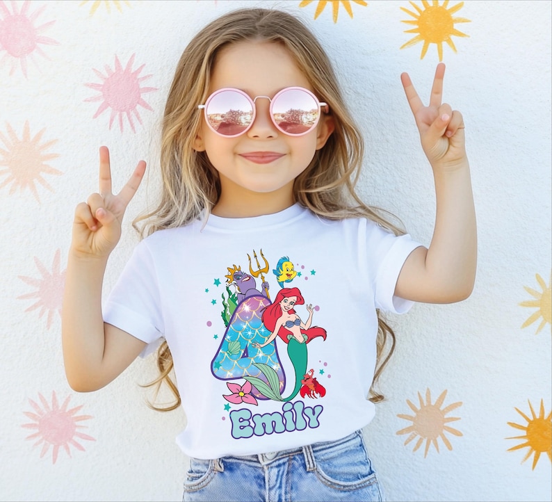A young girl wearing a white t-shirt with a mermaid design and sunglasses is making a peace sign with her hands.