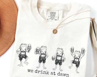May include: An off-white t-shirt with a black graphic print of four frogs wearing lederhosen and holding beer mugs. The text below the frogs reads "we drink at dawn". The shirt features a Comfort Colors brand label at the inner neckline.