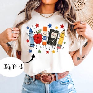 May include: A cream-colored t-shirt featuring a graphic print of five anthropomorphic school supplies including a blue gingham crayon, a red apple, an orange glue stick, a black polka-dot notebook, and a yellow pencil, surrounded by small stars.