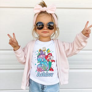 A young girl wearing sunglasses, a pink headband, and a white t-shirt with a mermaid graphic and the name "Ashley" printed on it, making a peace sign with her hands.