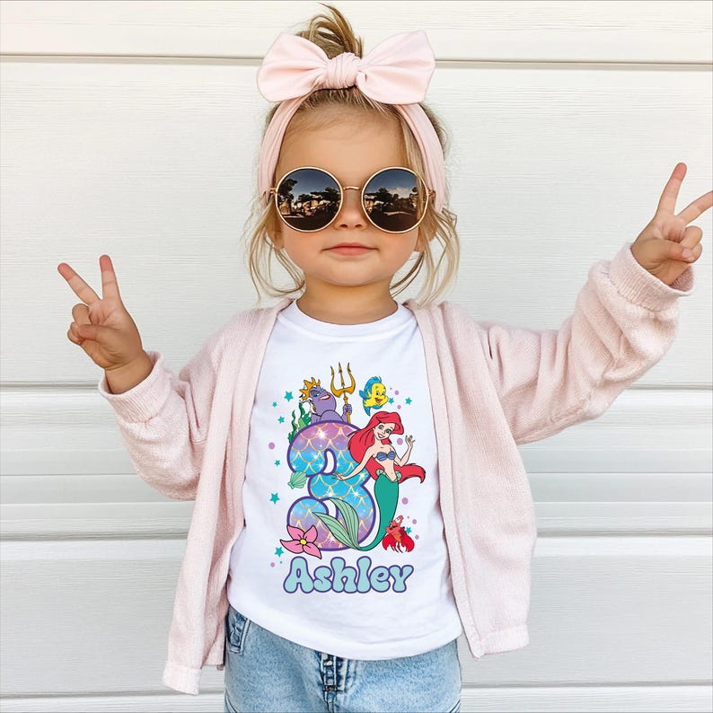 A young girl wearing sunglasses, a pink headband, and a white t-shirt with a mermaid graphic and the name "Ashley" printed on it, making a peace sign with her hands.