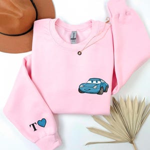 a pink sweatshirt with a blue car embroidered on it, along with a brown hat and some leaves.