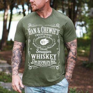 May include: Olive green t-shirt with a white graphic resembling a whiskey label. The label reads "Han & Chewie's Twelve Parsec Straight Smugglers Whiskey" with a spaceship illustration. The shirt also includes the text "70cl 40% Vol It May Not Look Like Much But It's Got It Where It Counts".