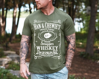 May include: Olive green t-shirt with a white graphic resembling a whiskey label. The label reads "Han & Chewie's Twelve Parsec Straight Smugglers Whiskey" with a spaceship illustration. The shirt also includes the text "70cl 40% Vol It May Not Look Like Much But It's Got It Where It Counts".