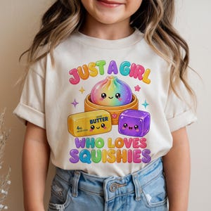 Könnte beinhalten: Ein cremefarbenes T-Shirt mit einer bunten Grafik und dem Text JUST A GIRL WHO LOVES SQUISHIES. Das Design zeigt einen regenbogenfarbenen Teigkloß in einem Bambusdämpfer, ein gelbes Stück gesalzene Butter mit der Aufschrift 4oz und einen lila Block.