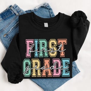 May include: Black sweatshirt with the words "FIRST GRADE" in colorful block letters. The word "first" is in pink, blue, yellow, and orange. The word "grade" is in green, orange, pink, and blue. The sweatshirt is folded on top of blue jeans.