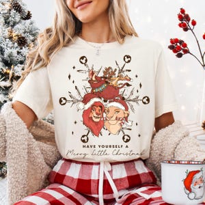 May include: Cream-colored t-shirt featuring a festive graphic of Lion King characters wearing Santa hats, set against a snowflake design. The text "Have Yourself a Merry Little Christmas" is printed below the graphic. The person is wearing red and white plaid pajama pants.