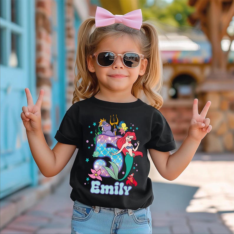 A young girl wearing a black t-shirt with a mermaid design and the name "Emilly" printed on it, smiling and making a peace sign with her hands.