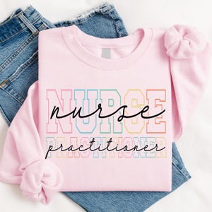 May include: A light pink sweatshirt with the words "NURSE PRACTITIONER" in a colorful, layered design. The word "NURSE" is in a bold, block letter style, with a cursive "nurse" overlay. The sweatshirt is folded over blue denim jeans.