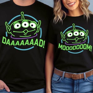 May include: Two black t-shirts with a green alien character graphic and neon text. The text on the first shirt says "DAAAAAAAAAD!" and the text on the second shirt says "MooooOOOOM!"