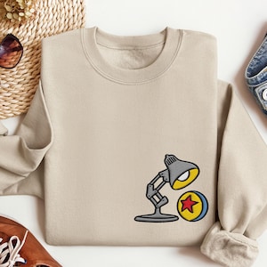 May include: Beige sweatshirt featuring an embroidered design of a gray desk lamp and a yellow, blue, and red ball. The sweatshirt has a crew neck and long sleeves. The design is on the lower right side of the shirt. The sweatshirt is made of a soft material.