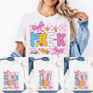 May include: White t-shirt with a colorful "PRE-K" graphic. The design includes a pink and blue checkered background, bows, pencils, and a lightbulb. The image also shows long-sleeved shirts with "Kindergarten", "First Grade", and "Second Grade" designs.