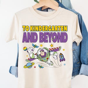 A white t-shirt with a colorful graphic design featuring a character from the Toy Story franchise, along with the text "TO KINDERGARTEN AND BEYOND" printed on it.