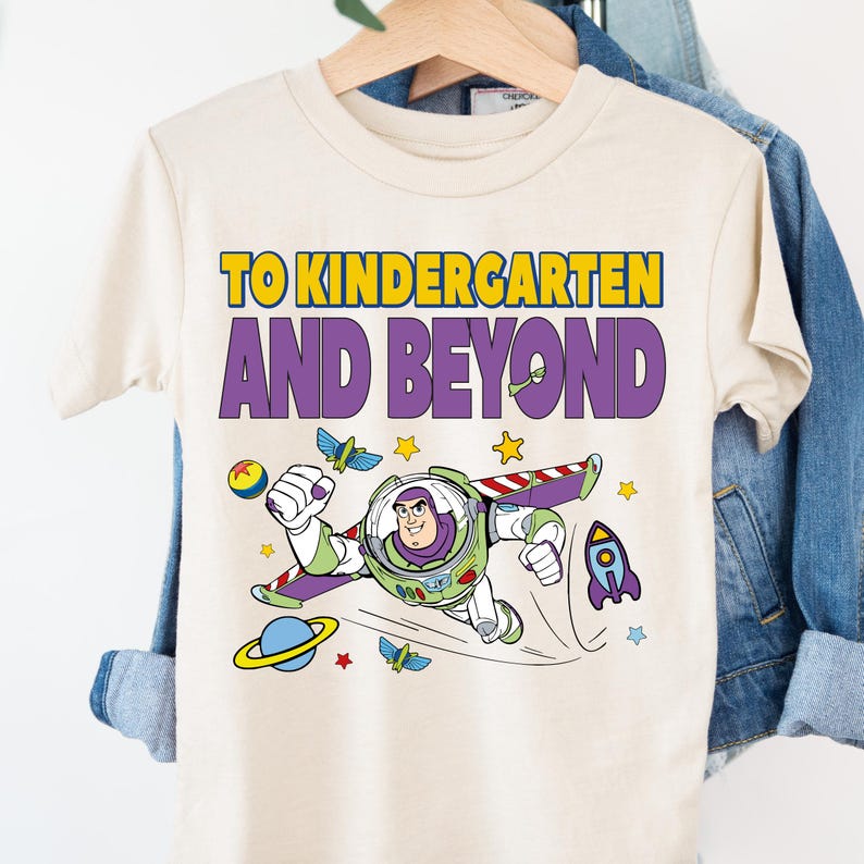 A white t-shirt with a colorful graphic design featuring a character from the Toy Story franchise, along with the text "TO KINDERGARTEN AND BEYOND" printed on it.