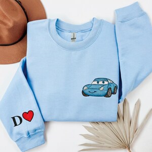 A light blue long-sleeved shirt with a blue car embroidered on it, along with a brown hat and a leaf, all laid out on a white background.