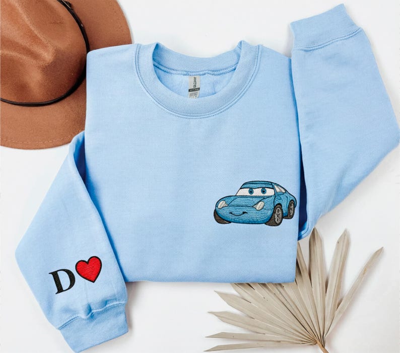 A light blue long-sleeved shirt with a blue car embroidered on it, along with a brown hat and a leaf, all laid out on a white background.